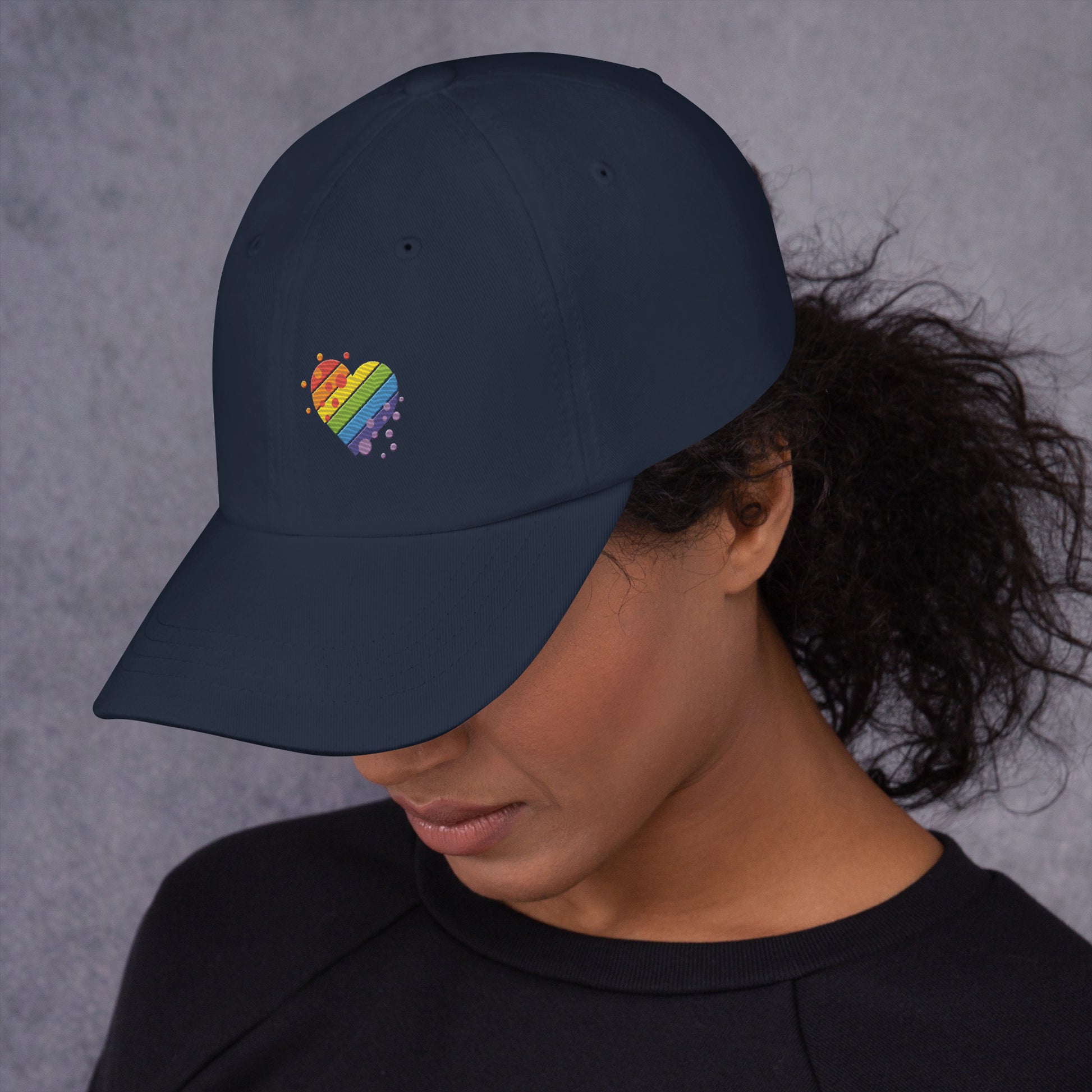 A female model wearing our navy baseball hat featuring rainbow heart design embroidery with a low profile, adjustable strap.