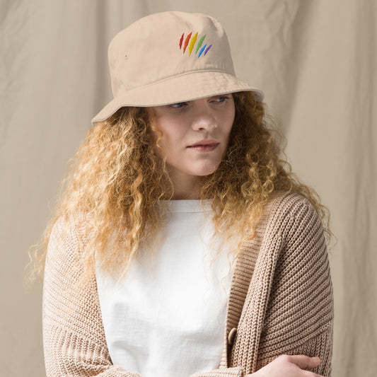 A female model sports a stone-colored bucket hat adorned with a rainbow-colored embroidered scratch mark on the front.