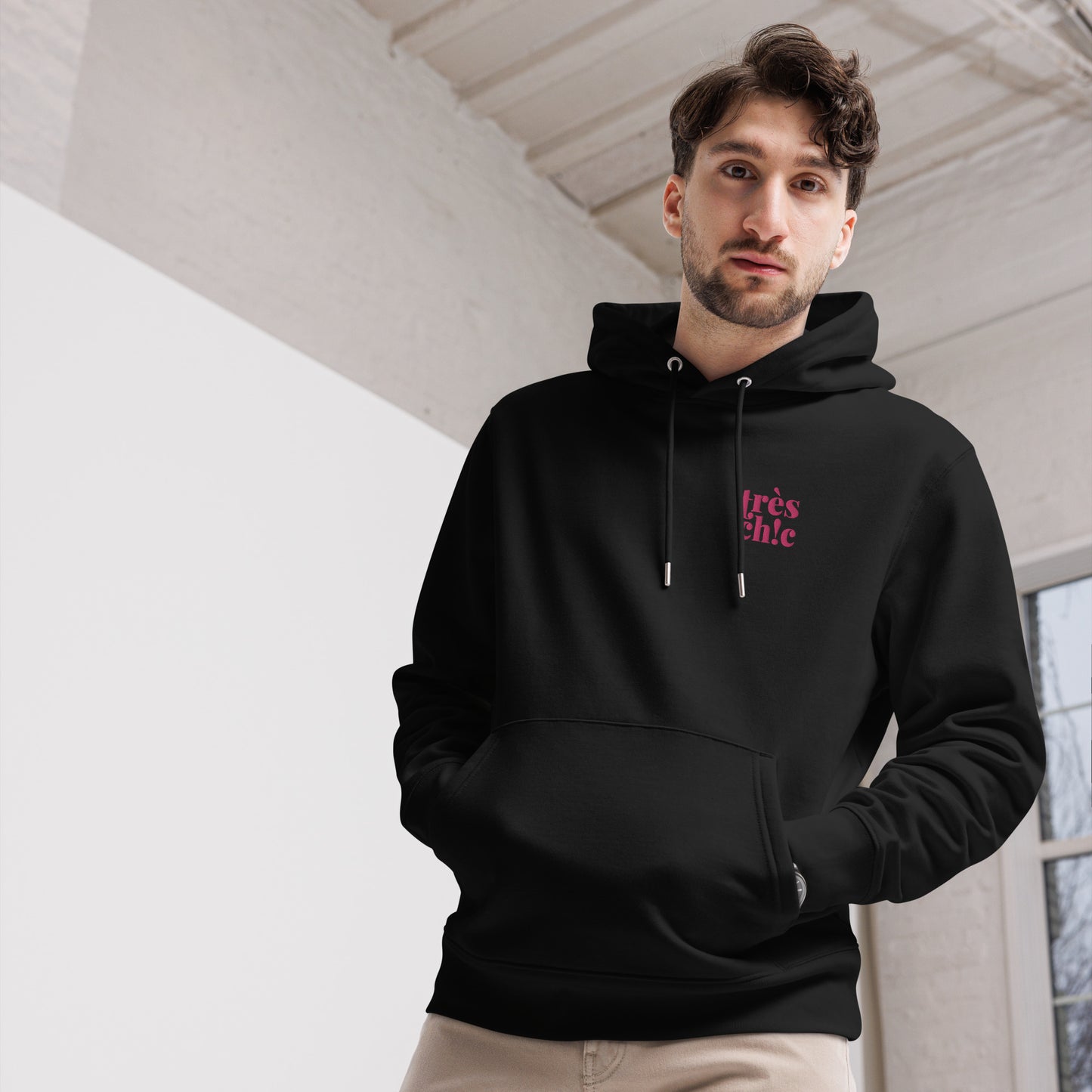 A male model wearing our unisex eco-friendly black hoodie featuring on the upper left chest; Trés Chic embroidery in hot pink color, adding a touch of lgbtq to your outfit. sizes: small, medium, large, extra large, double extra large.