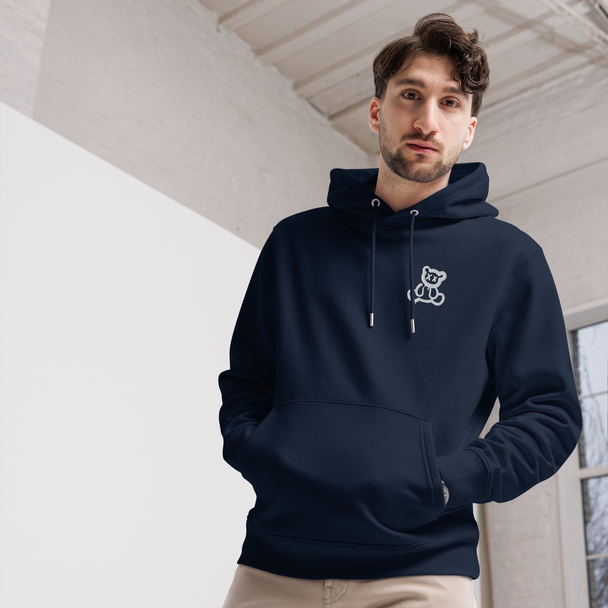A male model wearing our unisex eco-friendly french navy hoodie featuring on the upper left chest; a subtle embroidered graffiti bear in white, adding a touch of lgbtq to your outfit. sizes: small, medium, large, extra large, double extra large.
