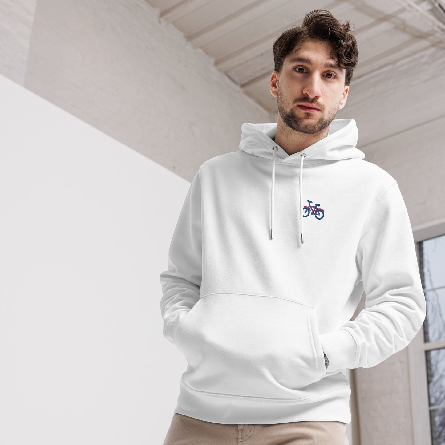 A male model wearing the unisex eco-friendly white hoodie featuring on the upper left chest; a subtle embroidered bicycle in bisexual colors, adding a touch of lgbtq to your outfit by putting the bi in bicycle. sizes: small, medium, large, extra large, double extra large.