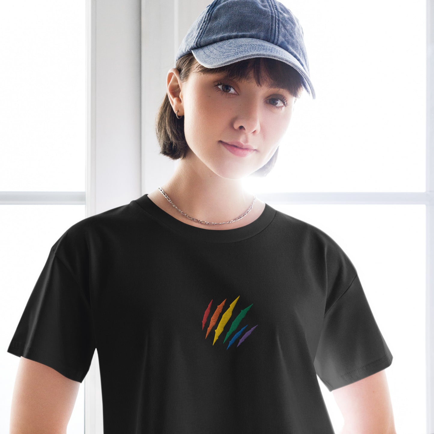 A female model wearing a relaxed, modest black crop top features a subtle rainbow mark embroidery. Adding a touch of rainbow to your look—a playful celebration of lgbtq culture! Made from 100% combed cotton. Available in Extra Small, Small, Medium, Large, Extra Large.
