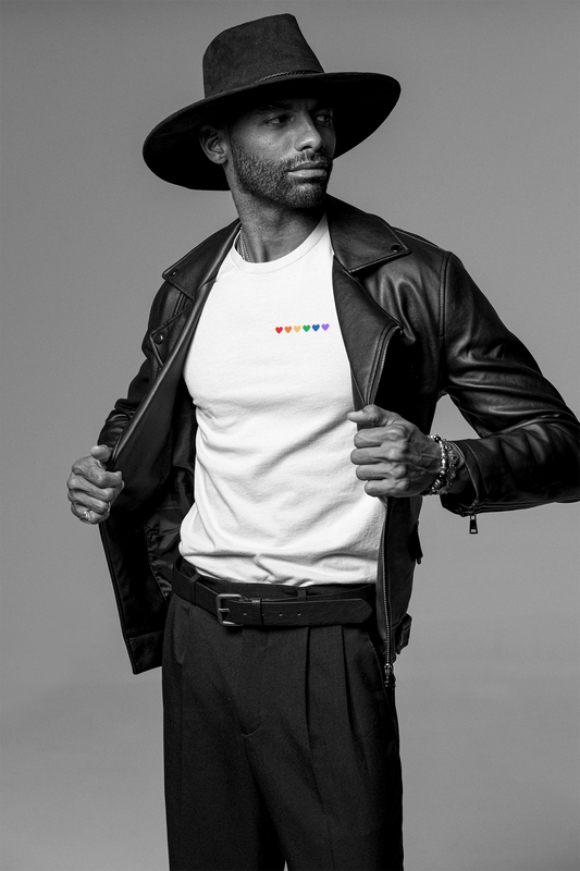 Male model wearing a fitted white organic cotton t-shirt with embroidered rainbow hearts on the left chest. Available in sizes S to 2XL.